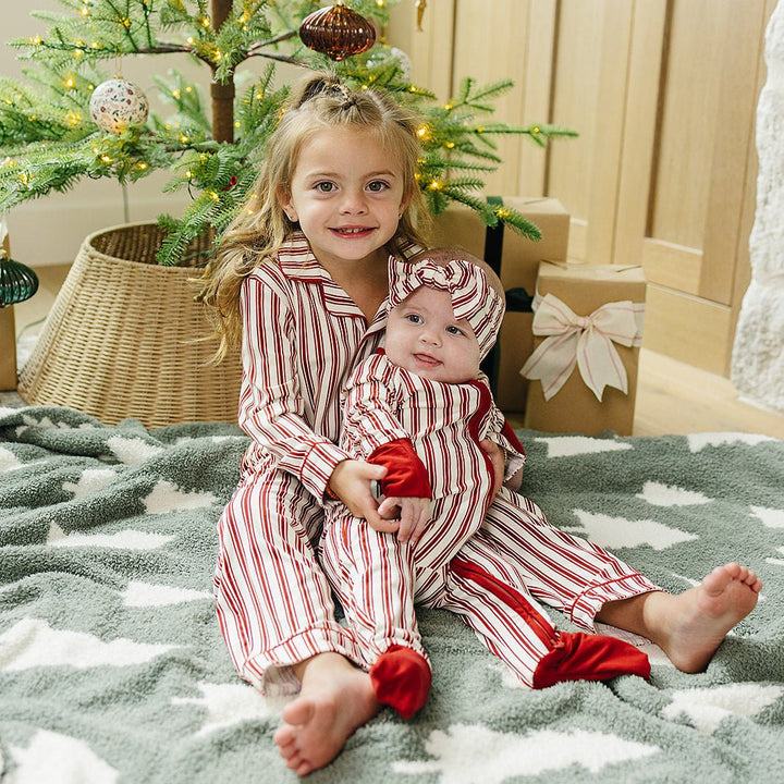 Two children in matching red and white pajamas sitting on a blanket in front of a decorated Christmas tree.