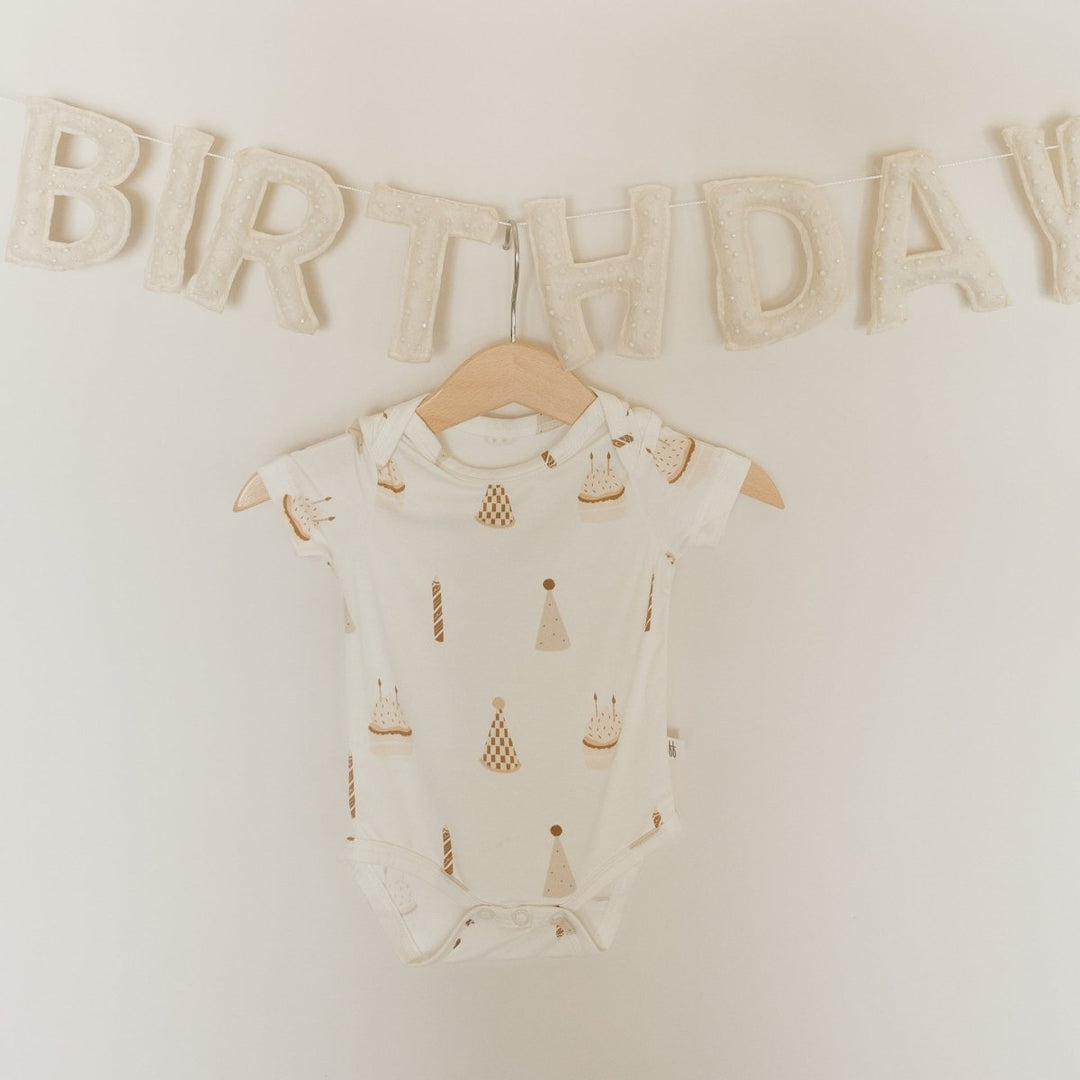 An off-white short-sleeve baby bodysuit with a gold and beige party hat and candle pattern, hanging on a wooden hanger against a cream wall with a &