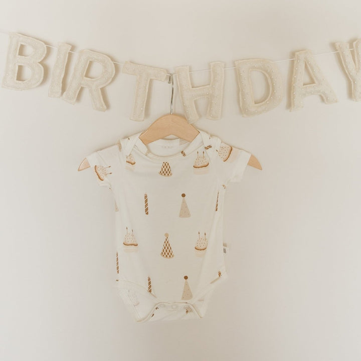 An off-white short-sleeve baby bodysuit with a gold and beige party hat and candle pattern, hanging on a wooden hanger against a cream wall with a &
