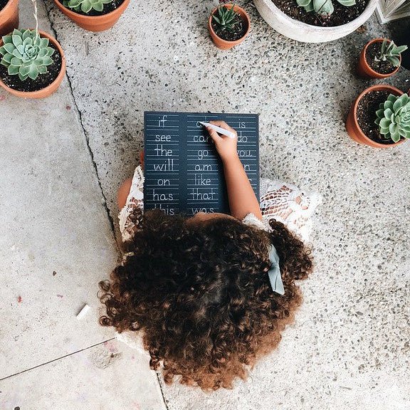 Overhead view of a person with curly brown hair sitting on a concrete floor surrounded by potted succulents, holding a small black chalkboard.