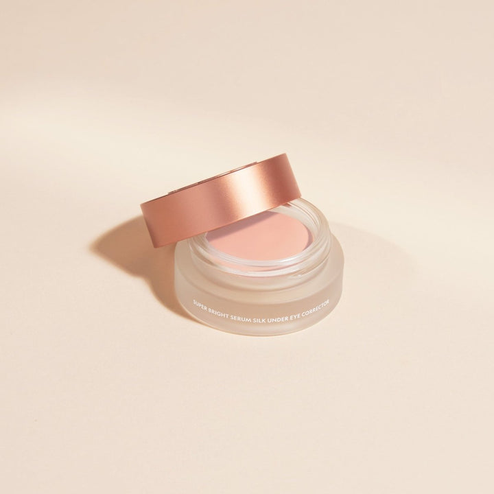 A small open cosmetic jar with a metallic rose-gold lid resting across the rim, showing a pale pink cream product inside. The jar is photographed on a clean warm beige studio background with a soft shadow.
