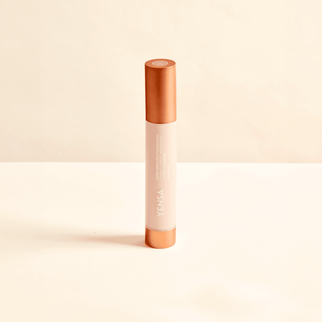 A single YENSA cosmetic tube standing upright on a clean, light beige surface against a matching beige background. The tube has a pale pink body with metallic rose gold caps at the top and bottom.