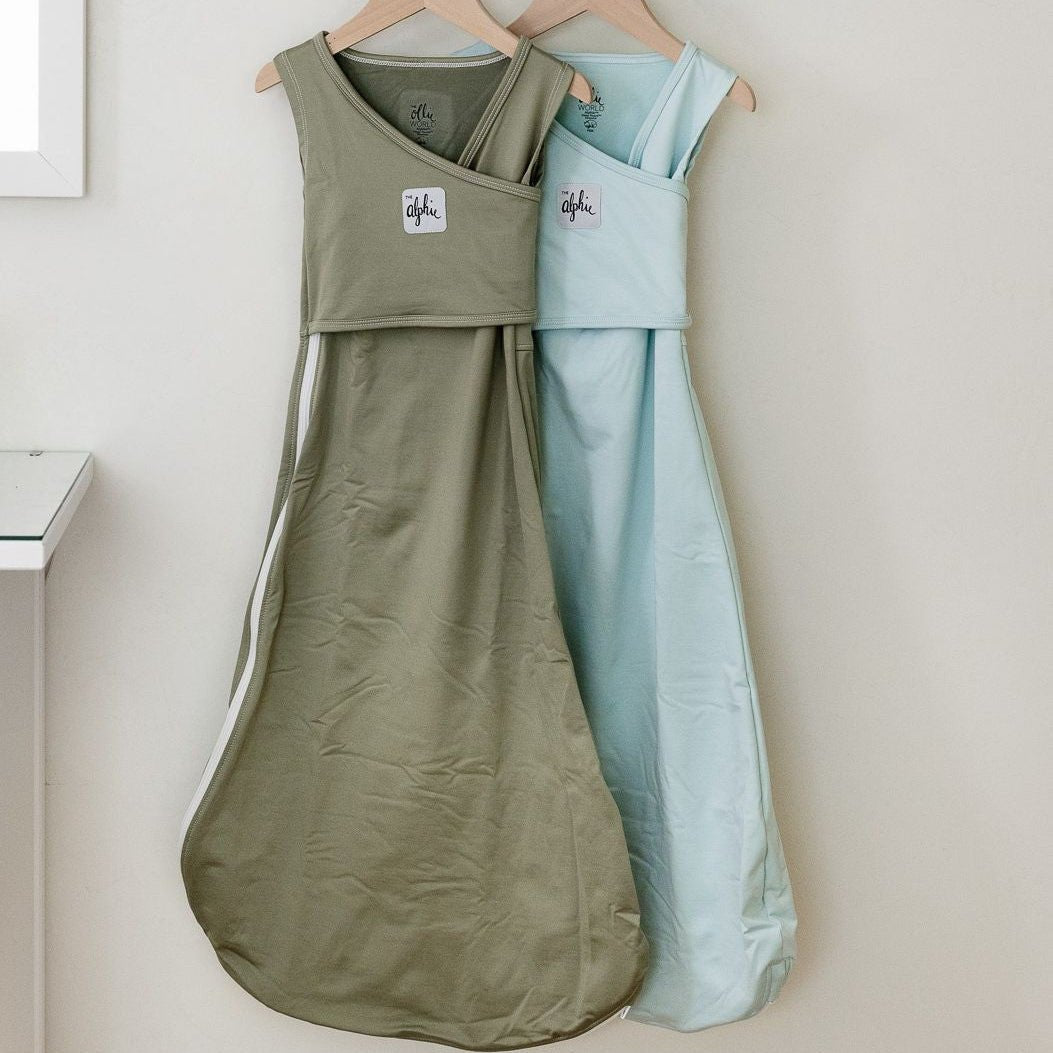 Two baby sleep bags hanging on wooden hangers against a light beige wall. The front sleep bag is olive green with a white side stripe and a small white logo patch, while the background sleep bag is light blue.