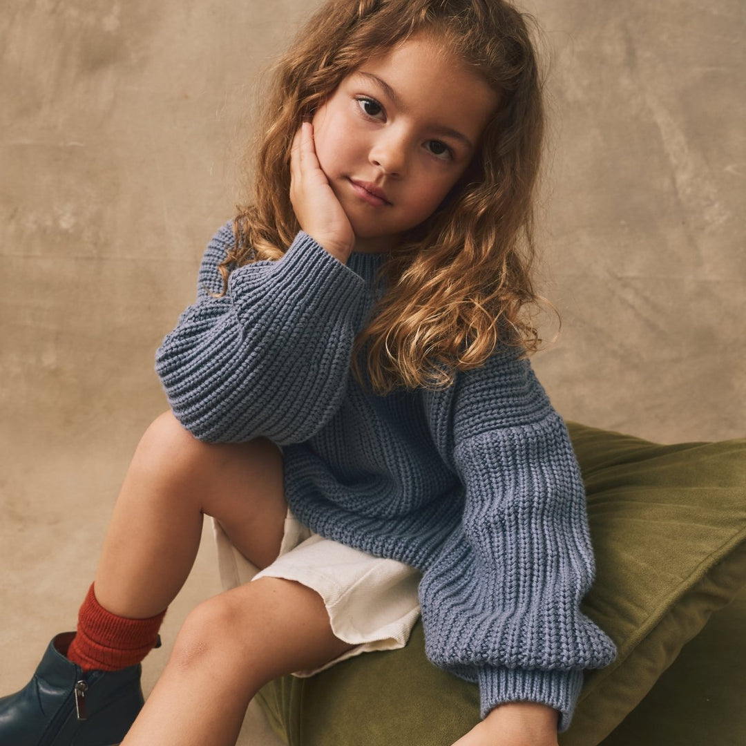 Young girl wearing a blue sweater and green pants sitting on a beige background
