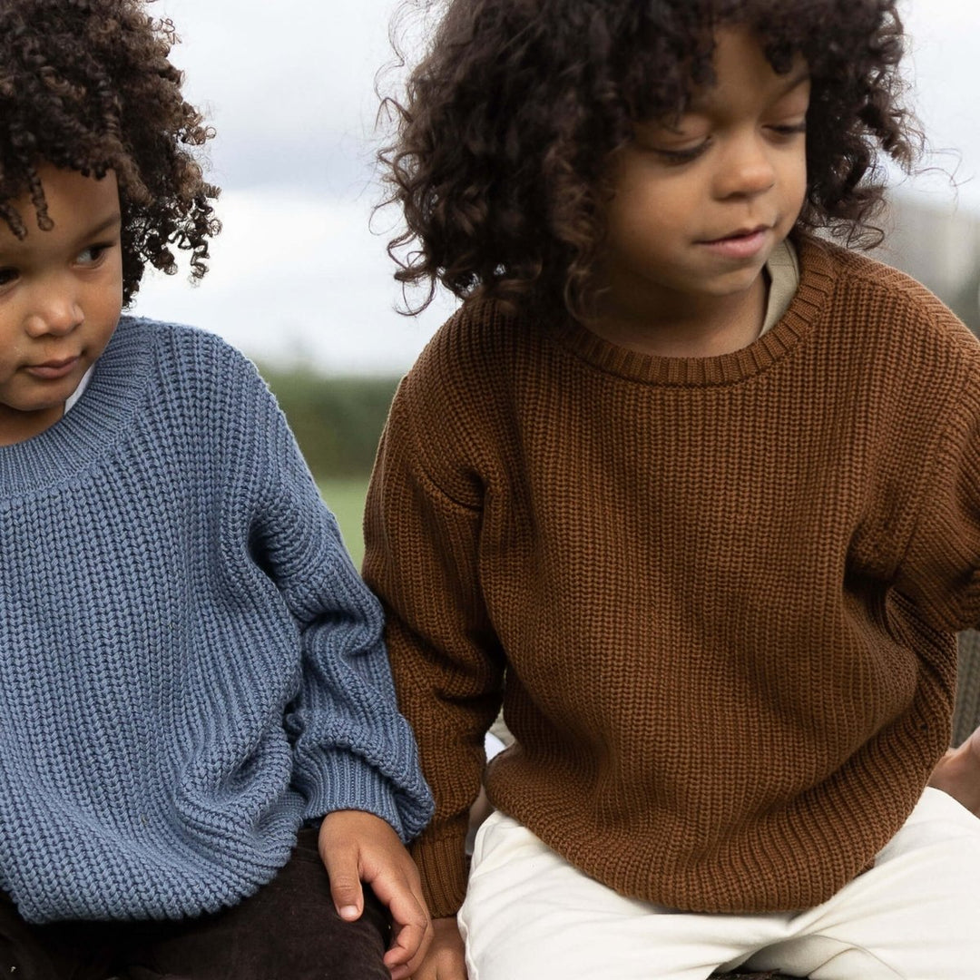 Two children sitting outdoors wearing knitted sweaters.