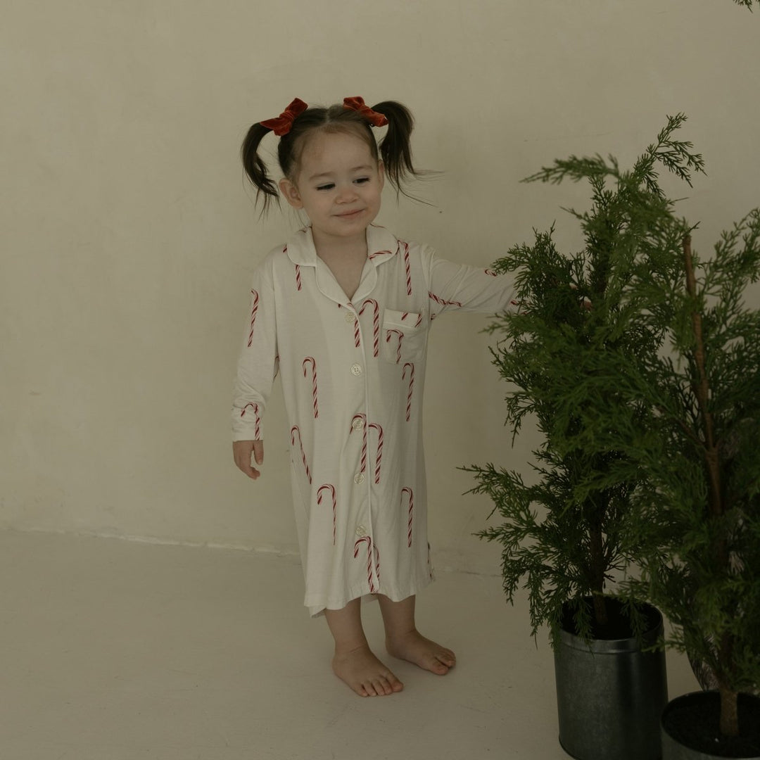 Young girl in a white dress with red patterns standing next to a potted plant in a room.