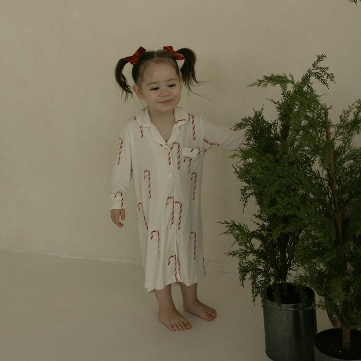 Young girl in a white dress with red patterns standing next to a potted plant in a room.