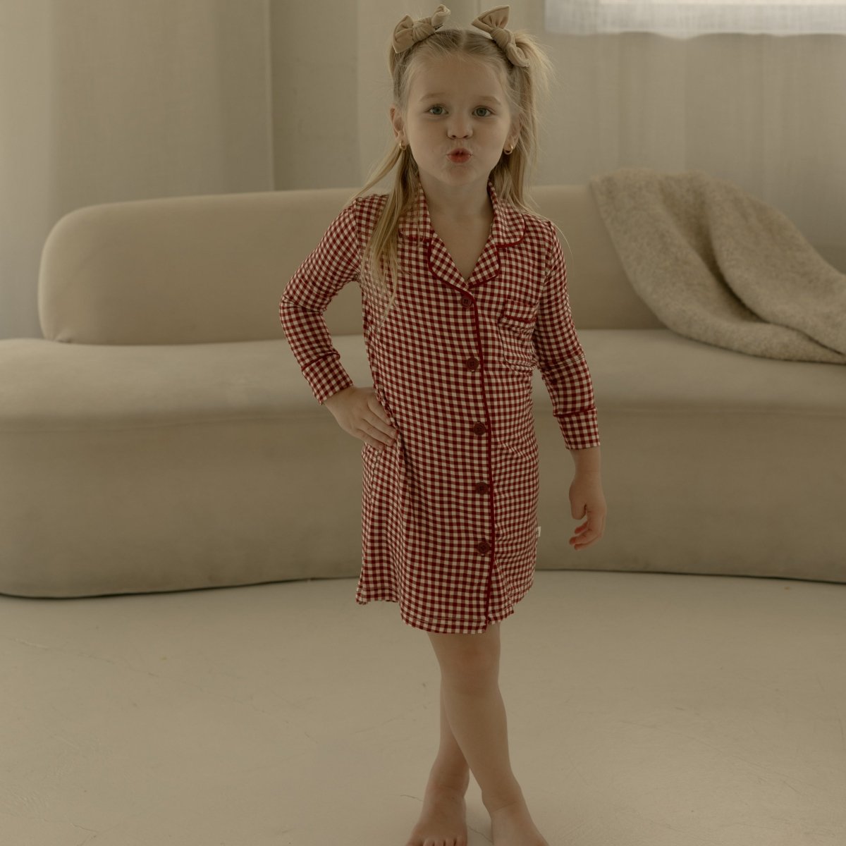 Young girl in a red checkered dress standing in a room with beige curtains and sofa.