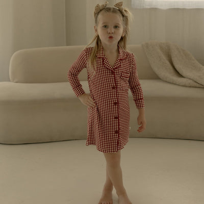 Young girl in a red checkered dress standing in a room with beige curtains and sofa.