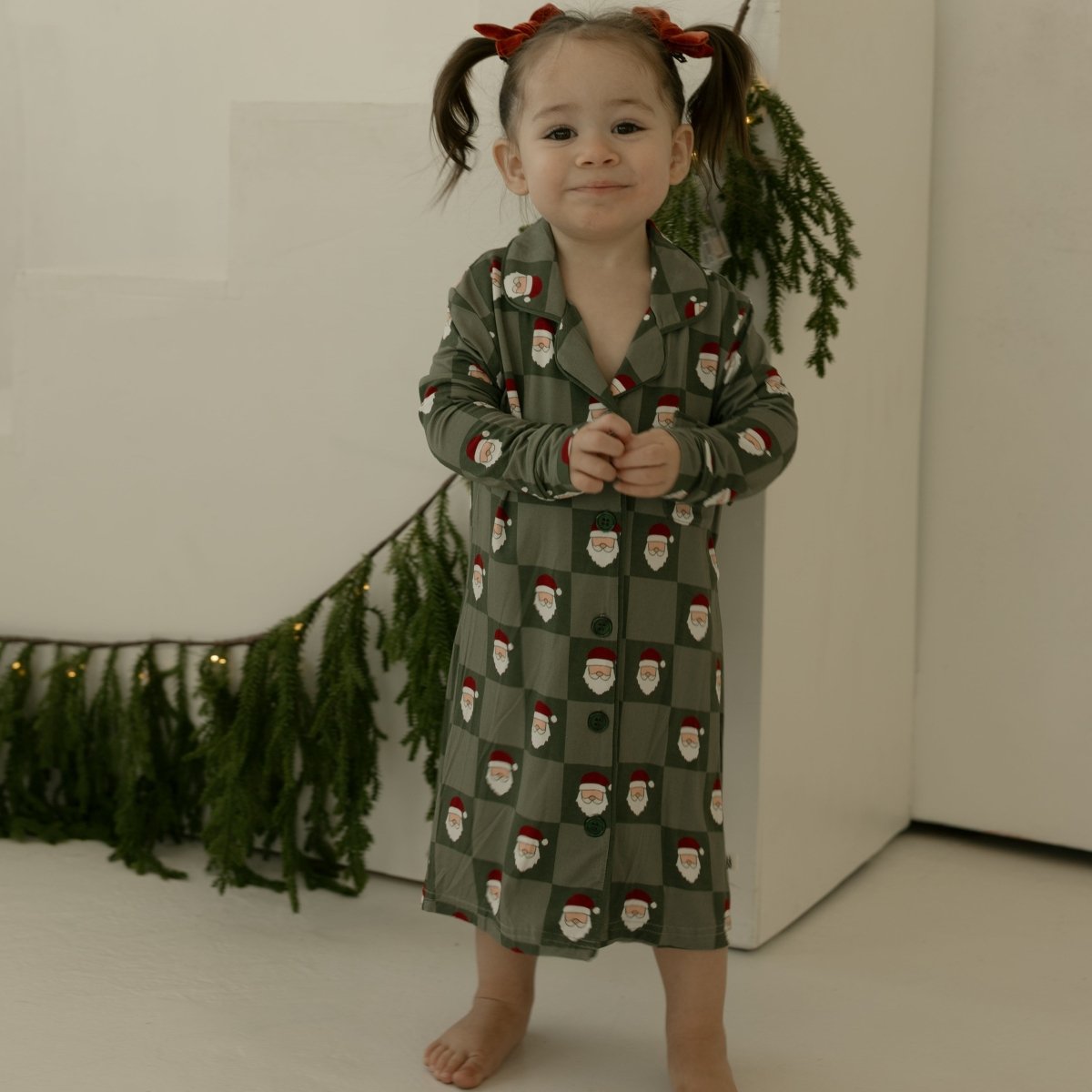 Child wearing a green pajama dress with Santa Claus pattern, standing against a white background.