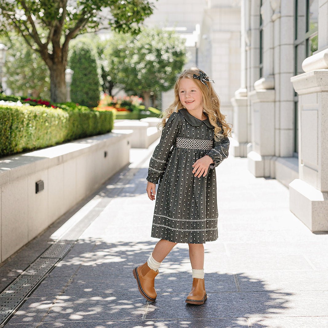 Young girl in a dress standing on a sunlit sidewalk with trees and a building in the background