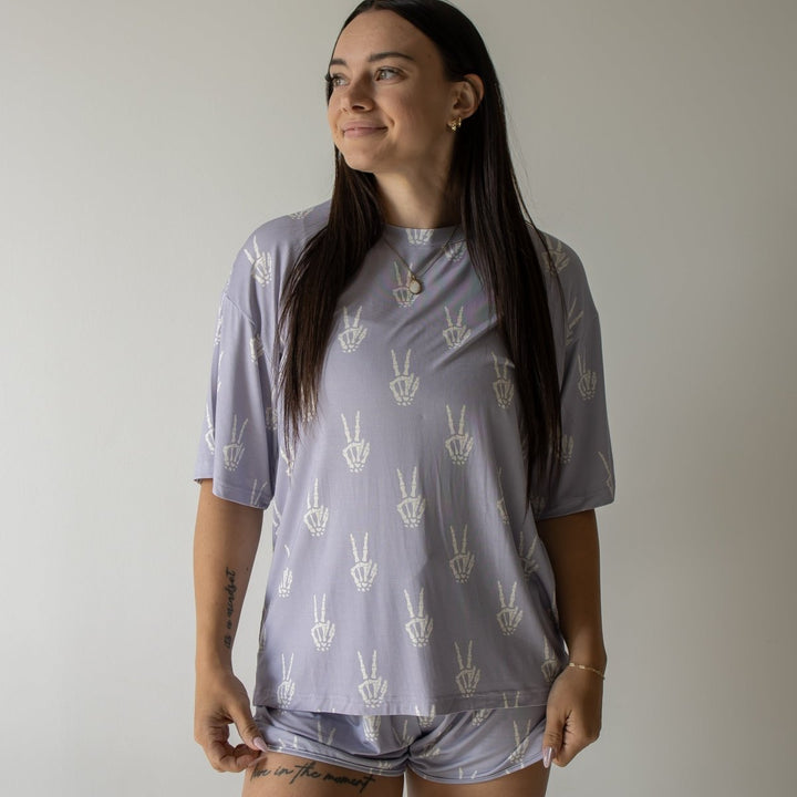 A woman modeling a matching lavender-gray short-sleeve t-shirt and shorts set with a white peace-sign pattern, standing against a plain light gray wall.